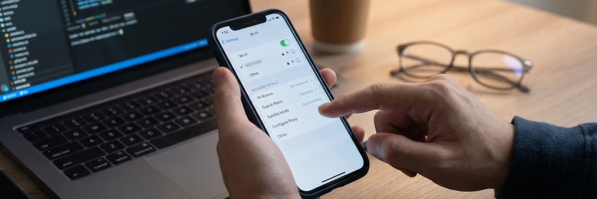 Человек настраивает Wi-Fi на смартфоне, рядом ноутбук и стакан кофе.