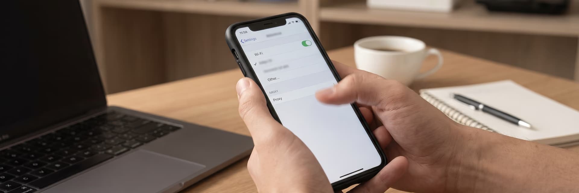 Человек настраивает Wi-Fi и прокси на смартфоне, сидя за столом с ноутбуком и чашкой кофе.